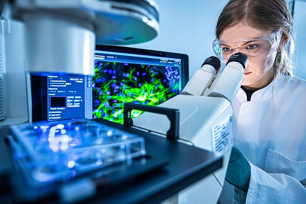 A scientist wearing safety goggles and a lab coat looks through a microscope. The microscope is focused on a sample slide illuminated by blue light. In the background, a computer monitor displays a detailed, colorful image of cells. The setting suggests a high-tech laboratory environment, focusing on cellular or microbiological research.