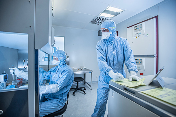 Two scientists work in full personal protective equipment. The scientist on the left of the image is manipulating materials with a micropipette in a biosafey cabinet while the scientist on the right observes and consults laboratory documents.