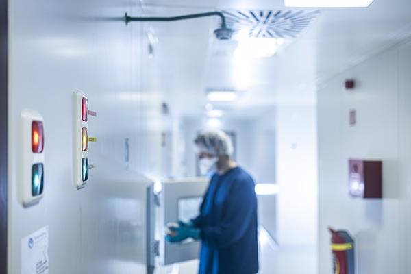 A blurred figure of a lab technician in a blue gown and face mask is seen working in the background of a sterile laboratory corridor. In the foreground, a wall-mounted panel with illuminated red, yellow, and green indicator lights is visible, suggesting a monitoring or alert system for the lab's equipment or processes. The lab environment is clean and well-lit, emphasizing a focus on safety and controlled conditions.